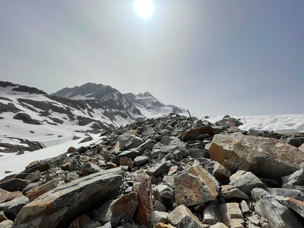 Die Seitenmoräne des Gaiskarferners, die Sonne scheint hell, der Gletscher ist tief verschneit und leuchtet weiß, der Weg übers Blockgelände ist gut mit Stangen markiert