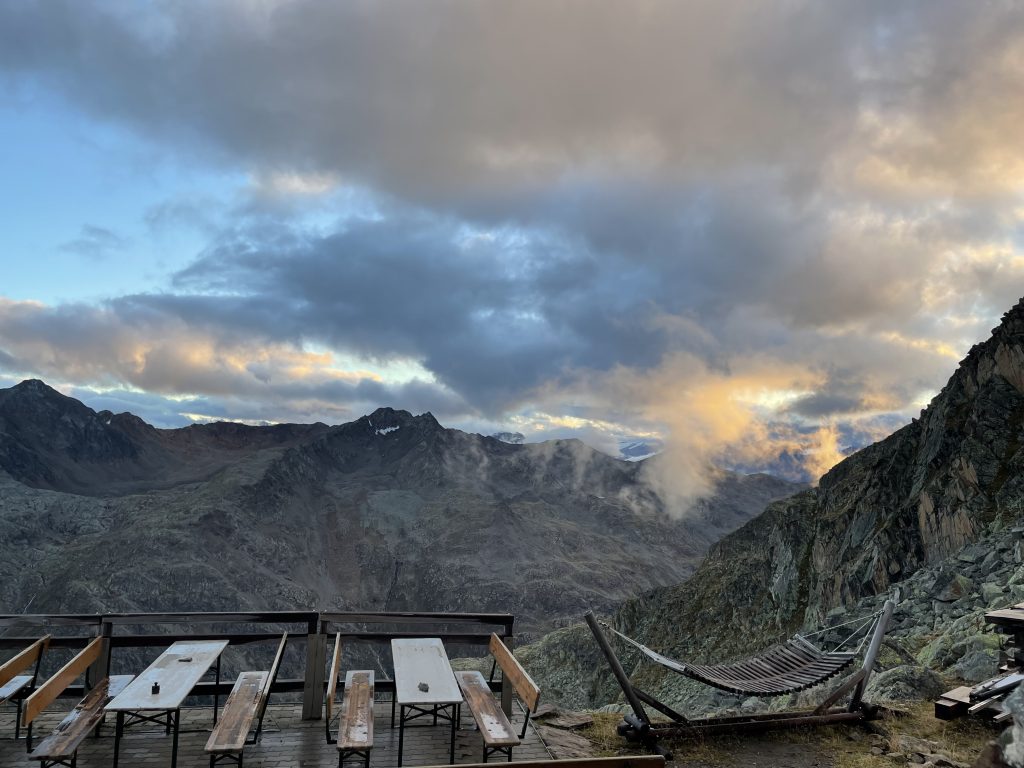 Die Terrasse der Hildesheimer Hütte mit Tischen bestuhlt, die Hängematte, sonnenbeleuchtete, tief hängende Wolken im Hintergrund, Stimmung nach einem Regenschauer