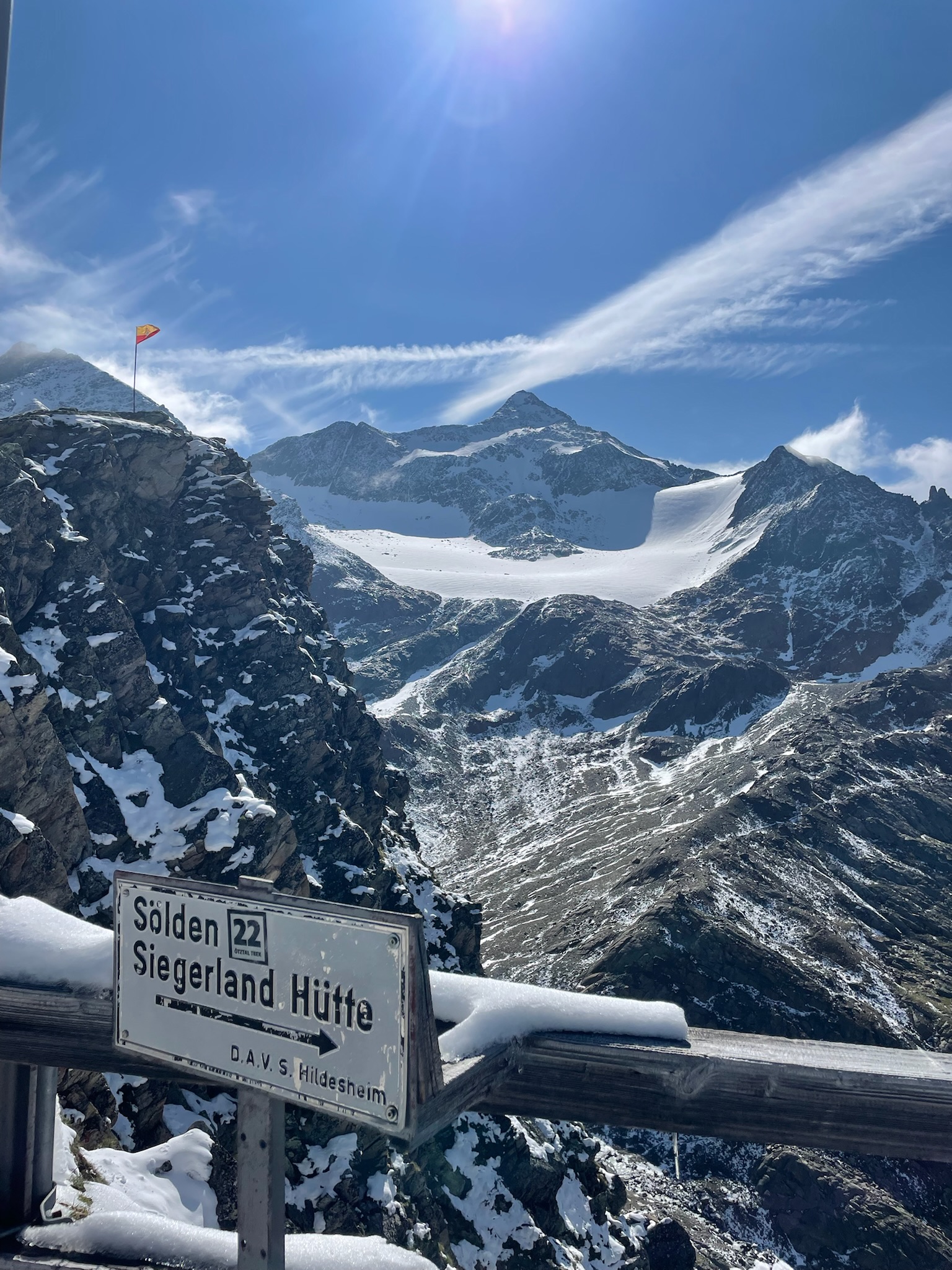 Blick von der Hütte zur Pfaffenschneid, der Neuschnee schmilzt in der warmen Sonne, der Gletscher glänzt weiß, die Fahne weht im Wind, im Vordergrund der Wegweiser zur Siegerlandhütte