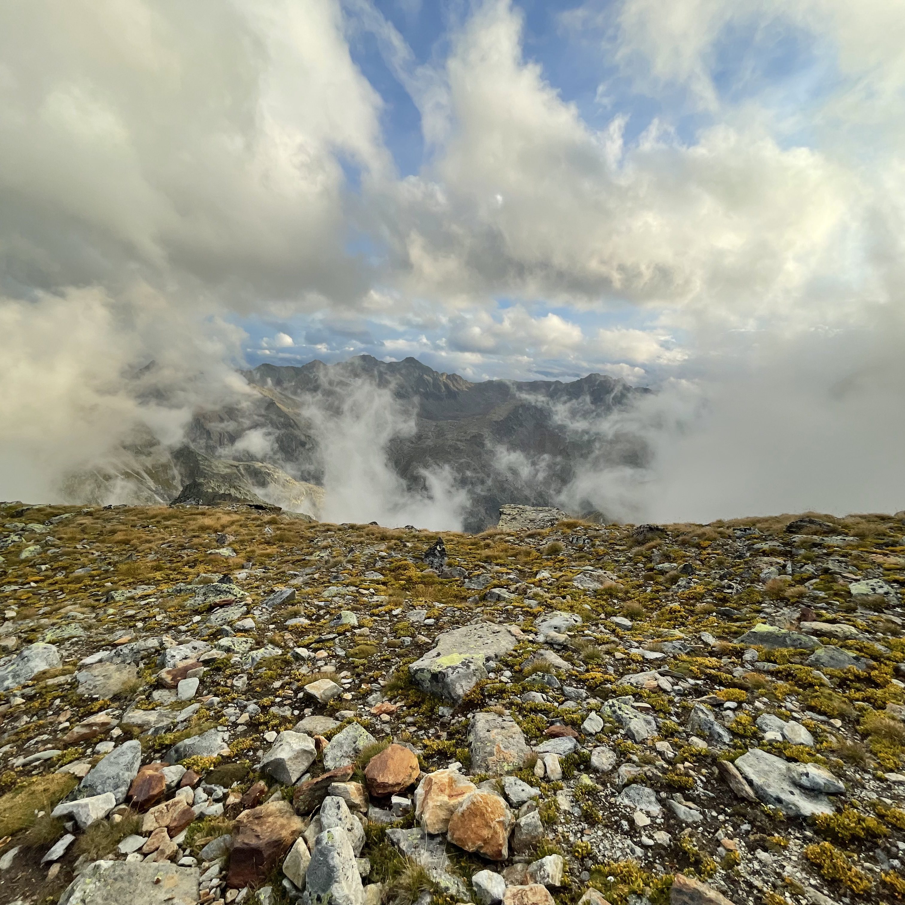 hochziehender Nebel mit Löchern, durch die das Bergpanorama sichtbar wird, im Vordergrund Felsen und gelb-grüne Wiese